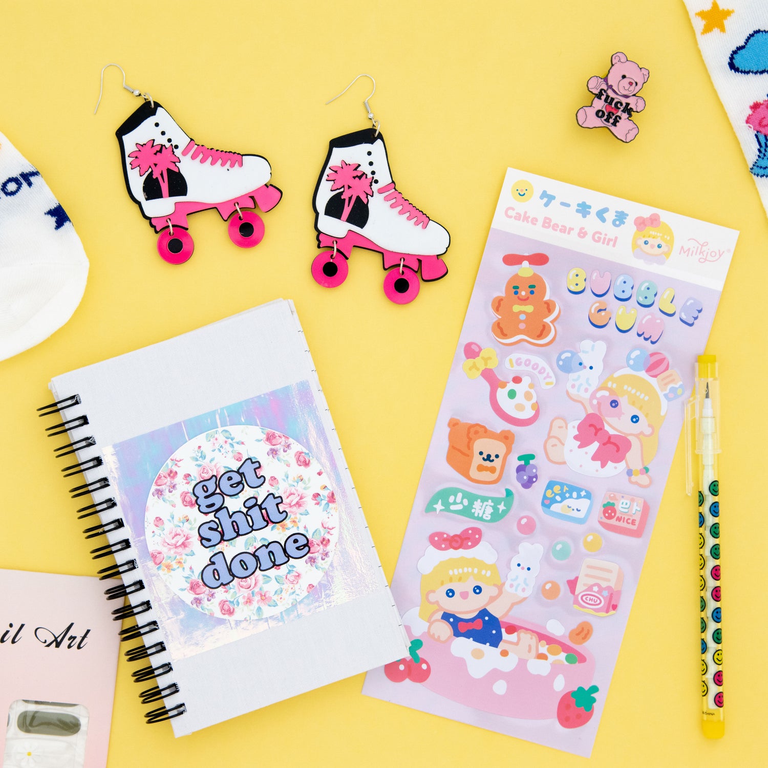 Funky roller skate earrings, enamel pin with a teddy bear saying 'Fuck Off,' bubble gum cake bear girl stickers, yellow smiley face pop a point pencil, and 'Get Shit Done' notebook with holographic sticker and flower background, perfect for fans of retro and quirky accessories from The Oddball Club.