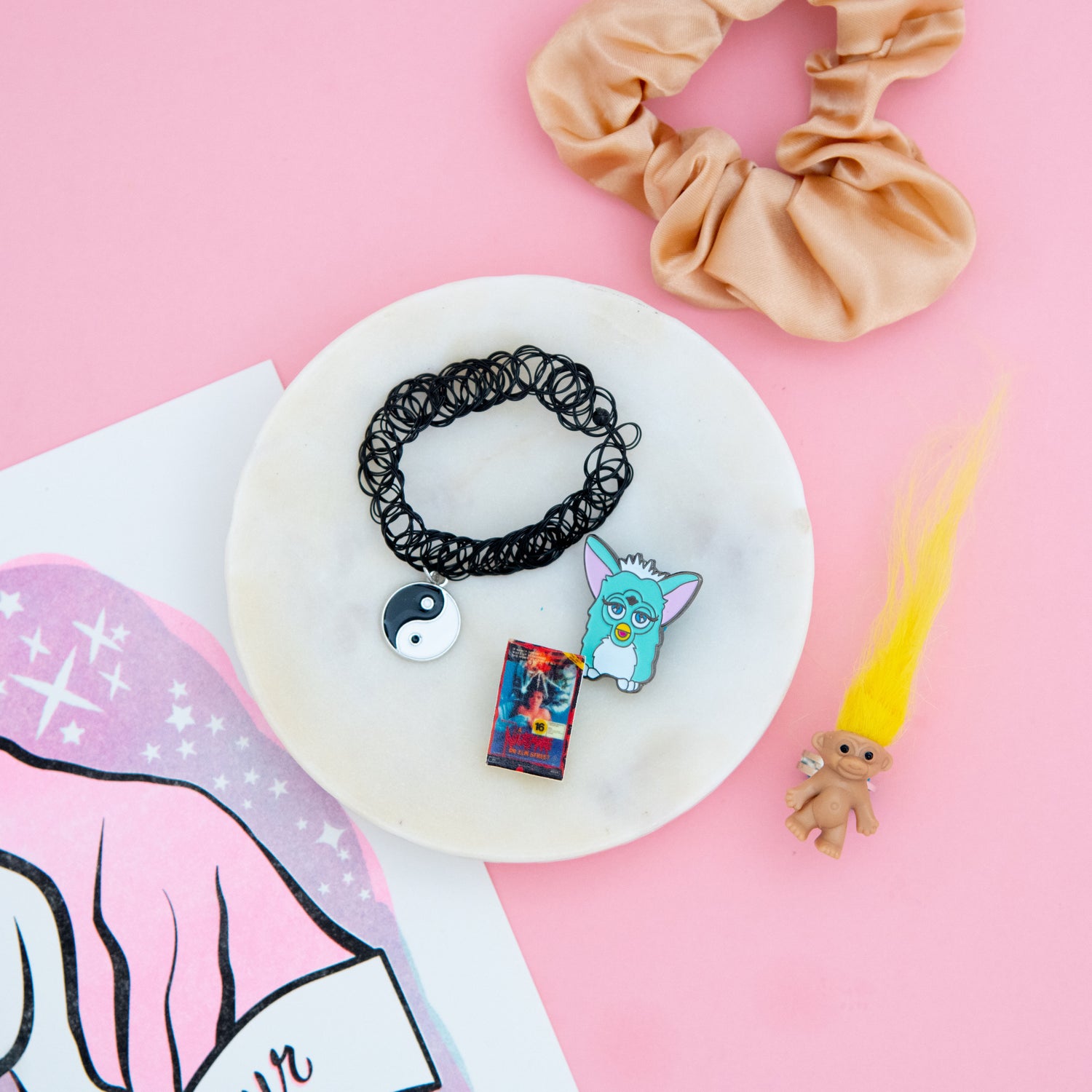 Get your '90s nostalgia fix with The Oddball Club subscription box! This month's box includes a trendy tan scrunchie, a stylish yin yang stretch necklace, a collectible Furby pin, a fun troll ring with yellow hair, all against a playful pink background. Join The Oddball Club today for unique and quirky finds!