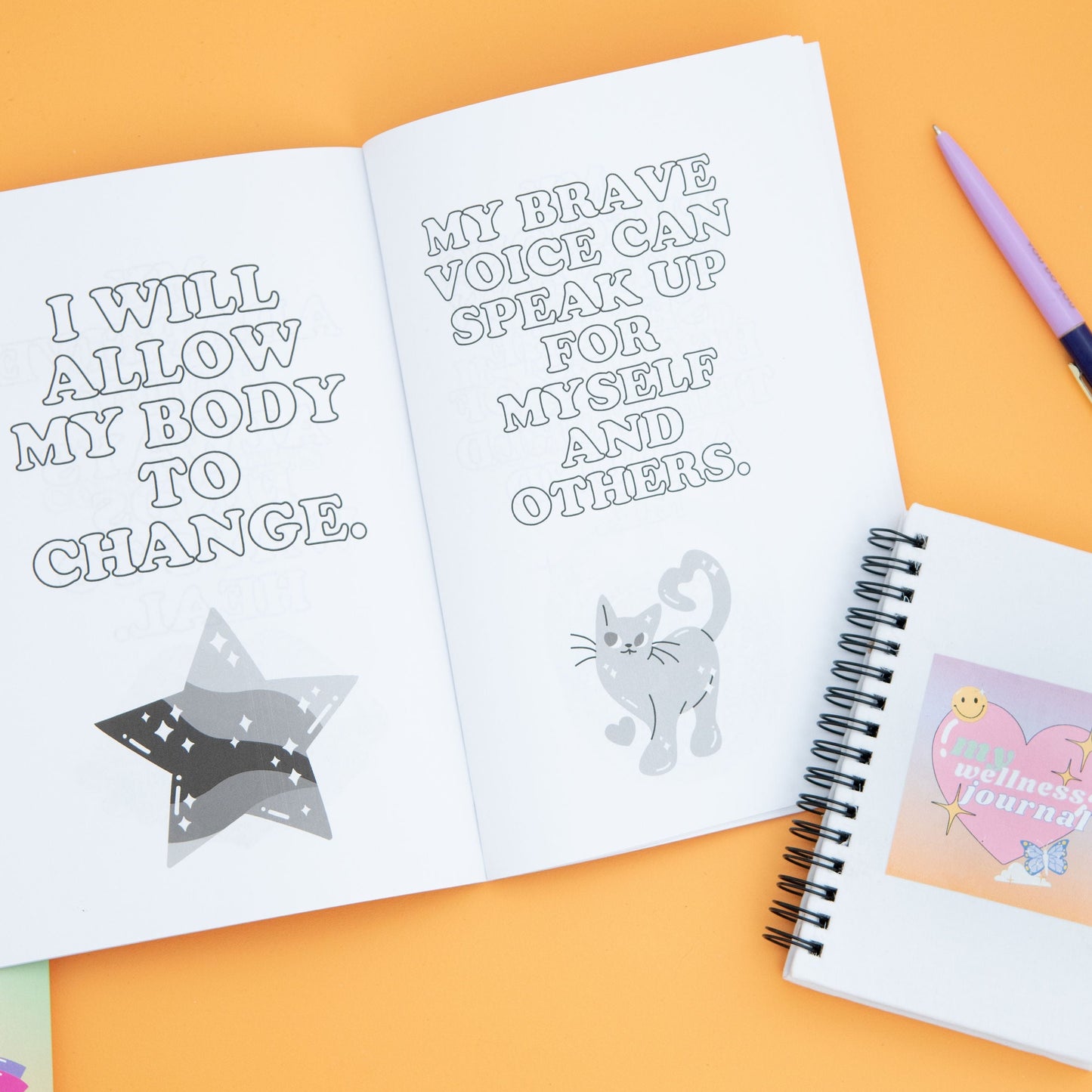 A positive body affirmation book with black and white star graphic on one page and a black and white cat on the other, next to a ballpoint pen and a spiral-bound white notebook with heart art cover labeled 'My Wellness Journal.' The perfect stationary for practicing body positivity and self-care.