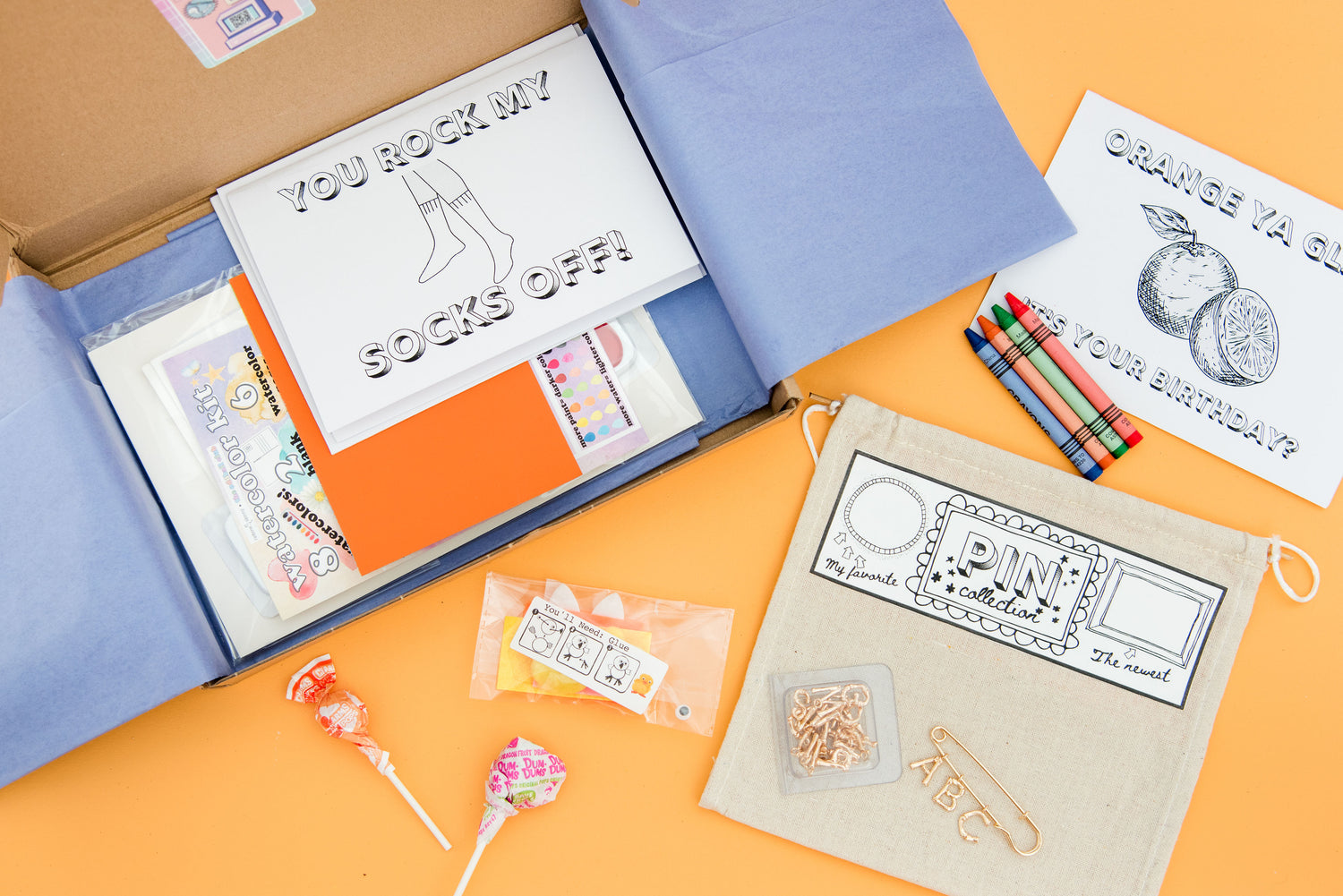 An Oddball Club DIY-themed subscription box opened with purple tissue paper. Box contents include a greeting card with outlined items for coloring, a set of crayons, a set of watercolor art kit, lollipops, chick DIY craft, a pink banner, and a DIY alphabet pin.