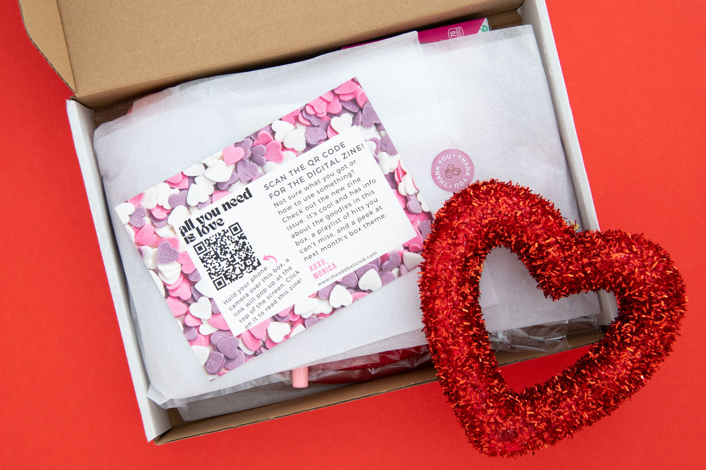 Love-themed subscription box contents with red sparkle heart on white tissue paper - heart-shaped sunglasses, rose petal soap, heart-shaped candle, love potion bath bombs, heart-shaped silicone ice cube tray, and love-themed greeting card with roses.