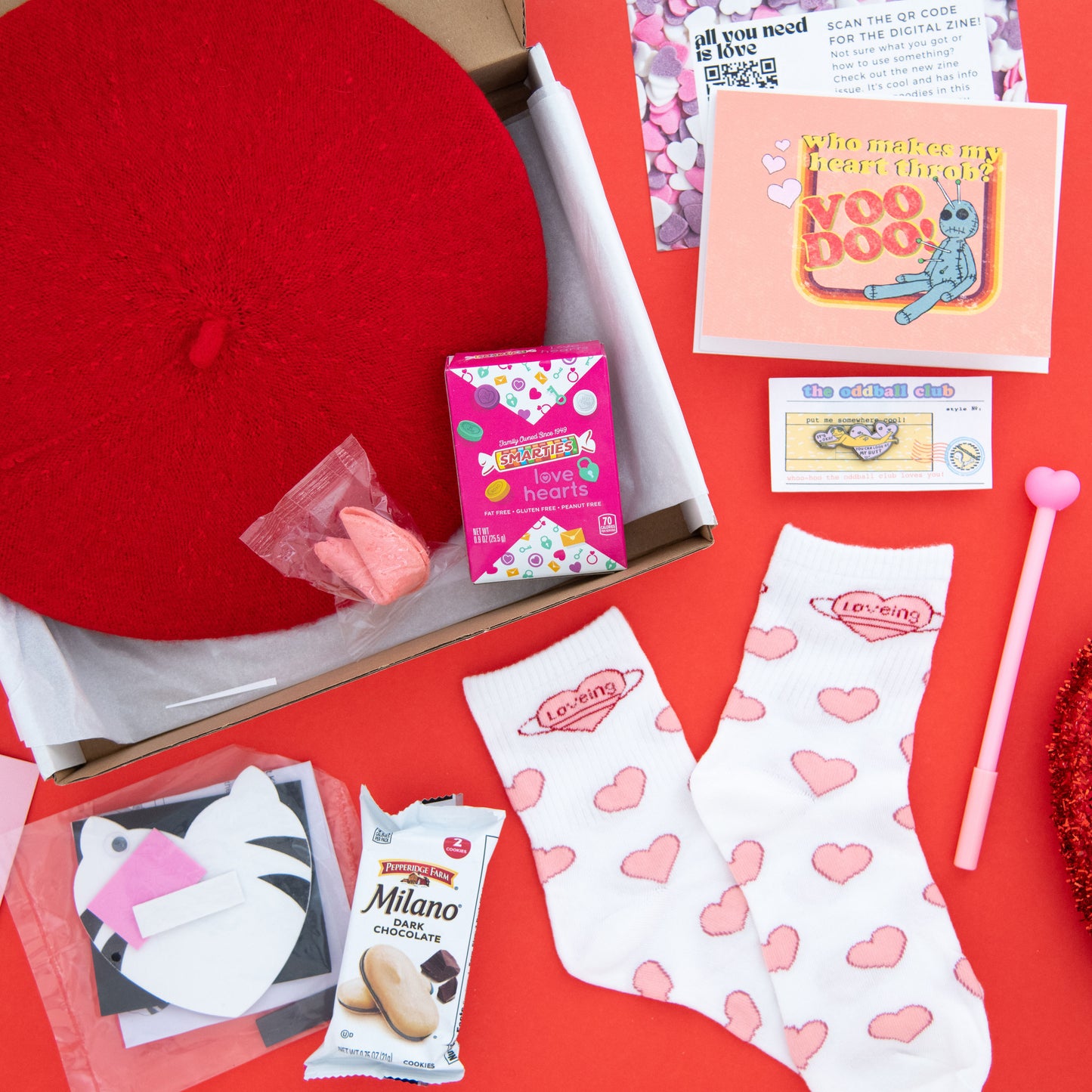 Heart-patterned socks with loving words, Smarties conversation heart candies box, pink fortune cookie, peach voodoo greeting card with a voodoo doll with pins, enamel pin, red French beret, heart pen, and a French beret subscription box
