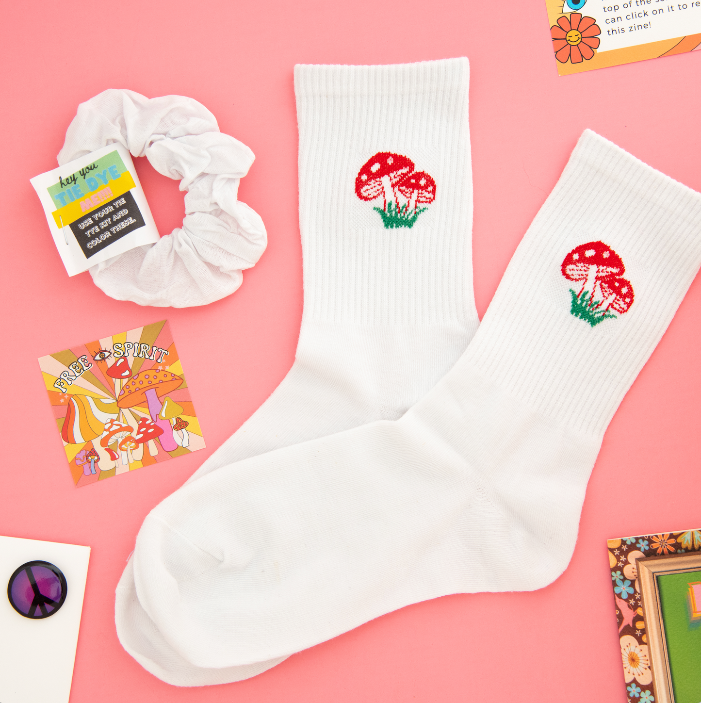 Hippie vibe subscription box with white socks featuring red mushrooms and green grass, free spirit square sticker, purple peace sign pin, and white scrunchie. Perfect for bohemian and free-spirited individuals