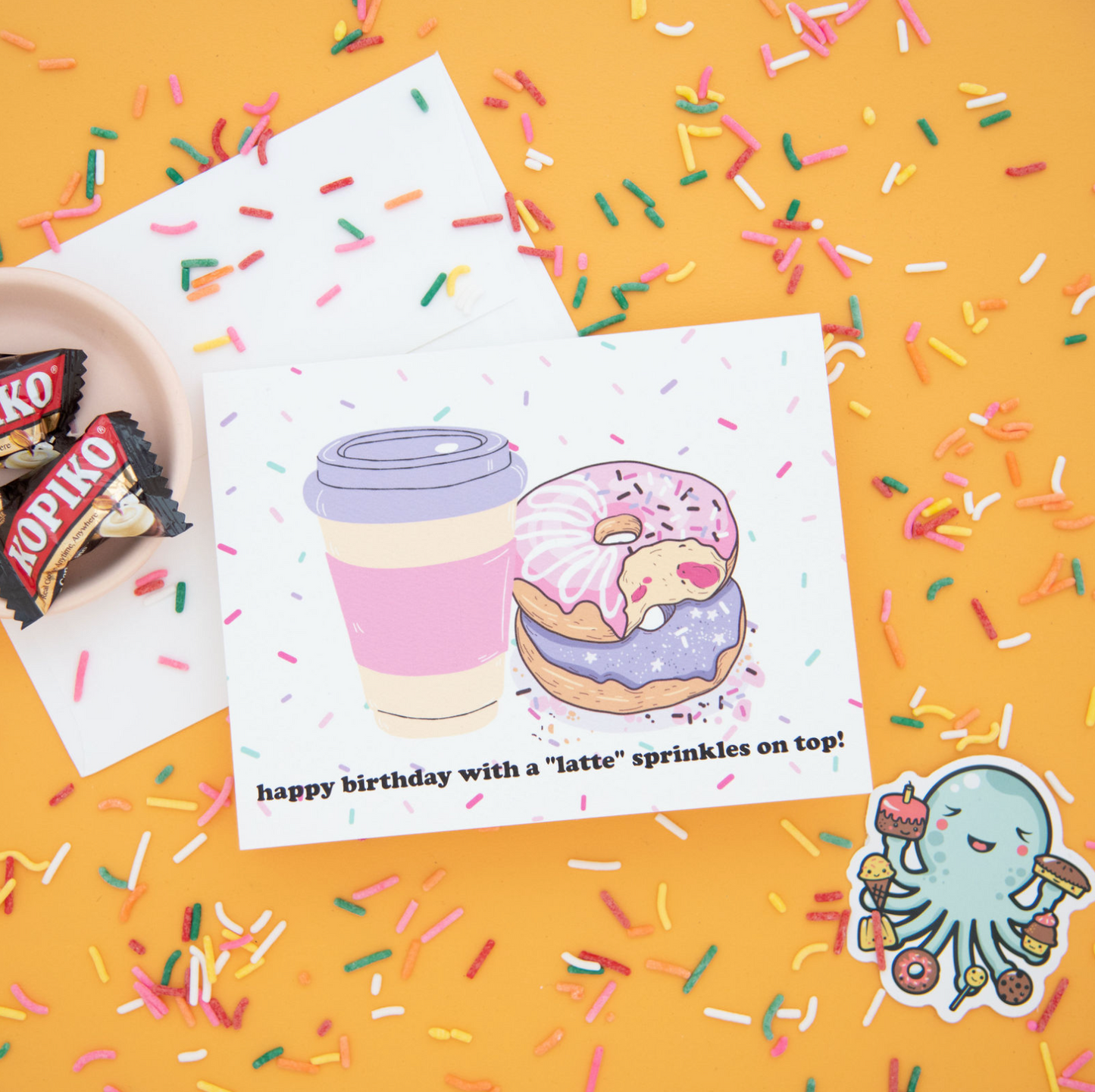 A birthday-themed greeting card with a coffee cup and two donuts stacked and sprinkles on top, surrounded by colorful sprinkles, along with an octopus sticker featuring donuts in the corner. Also included in the image is a pack of Kopiko candy. The image is set against a bright yellow background.